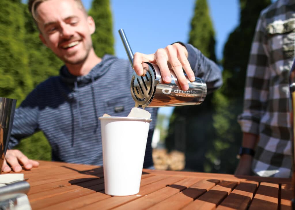 Person pouring mixed beverage from a branded cocktail shaker at an outdoor event.